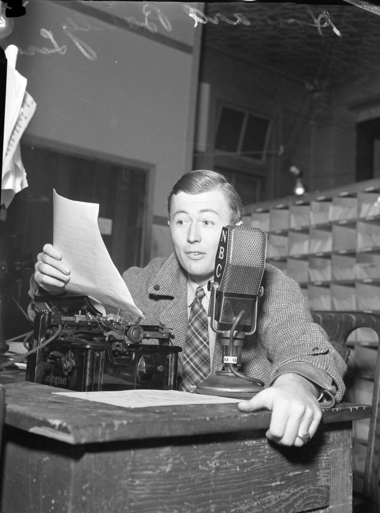 Black and white photo of Newscaster Howard Bailey reading off a piece of paper and sitting in front of an NBC microphone