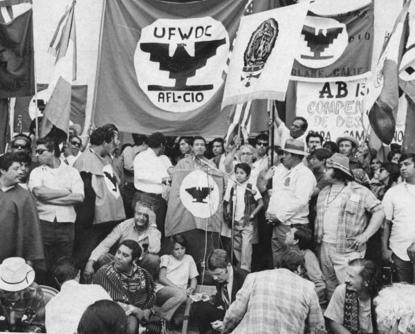 Cesar Chavez speaking to a large audience with the United Farm Workers flag behind him.