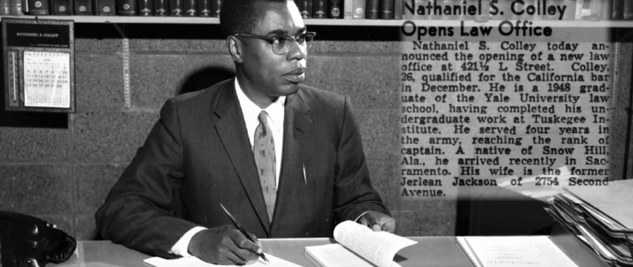 Black and white photo of Nathaniel Colley at his desk with a newspaper clipping shown next to him