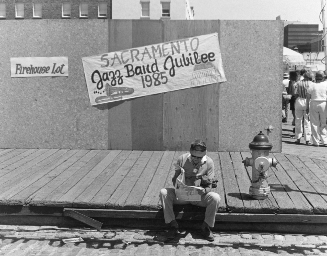 A man reads a program sitting on the sidewalk with a banner reading, "Sacramento Jazz Band Jubilee, 1985" hangs at a tilt behind him.