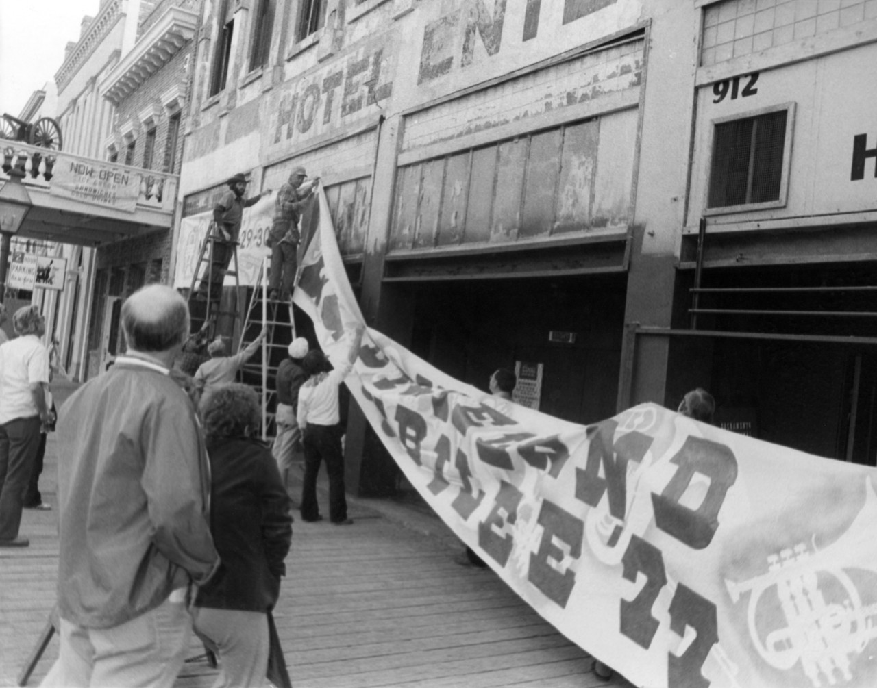 A group of men, two on ladders, under the Hotel Enterprise, hanging a large Jazz Jubilee banner.