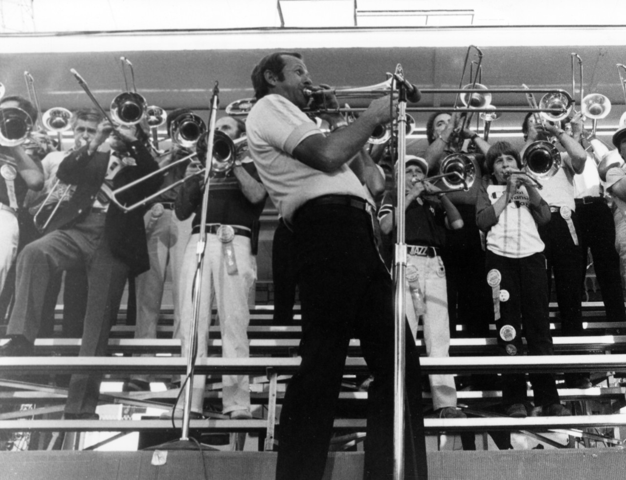 A crowd of trombone players are standing in stadium seats. One man is playing into a microphone in the middle.