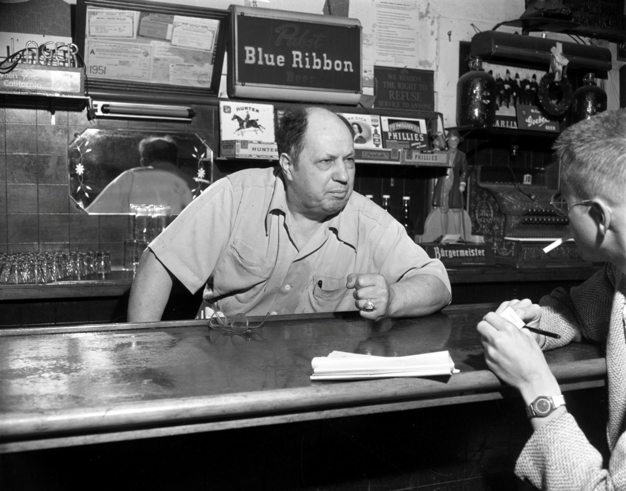 A bartender slams his fist on the bar while talking to a patron seated across from him