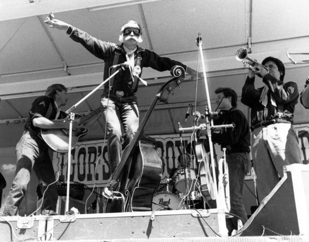 A man rides a standing bass on stage for a performance. Three other men are on stage with him and they are surrounded by equipment.