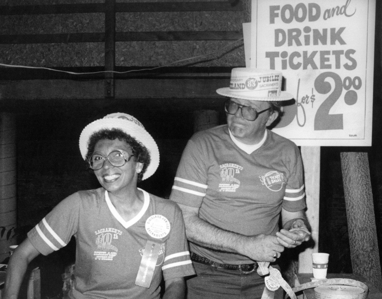A man and a woman volunteer with Jubilee T-Shirts smile for the camera with a sign behind them reading, "Food and Drink Tickets for $2.00"